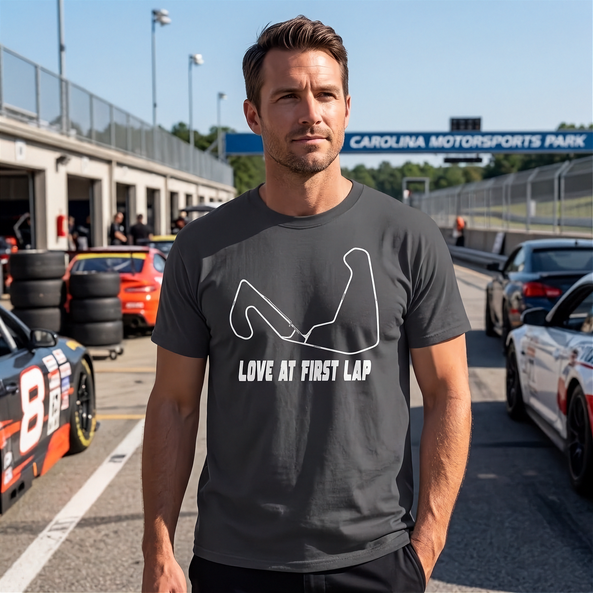 A man standing on a race track pit lane wearing a dark gray t-shirt with a white graphic of a racing lap and the text 'LOVE AT FIRST LAP'.
