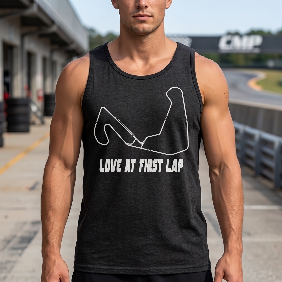A man wearing a dark charcoal gray sleeveless tank top with a white graphic of a racing arm and the text 'LOVE AT FIRST LAP' stands on a racetrack.