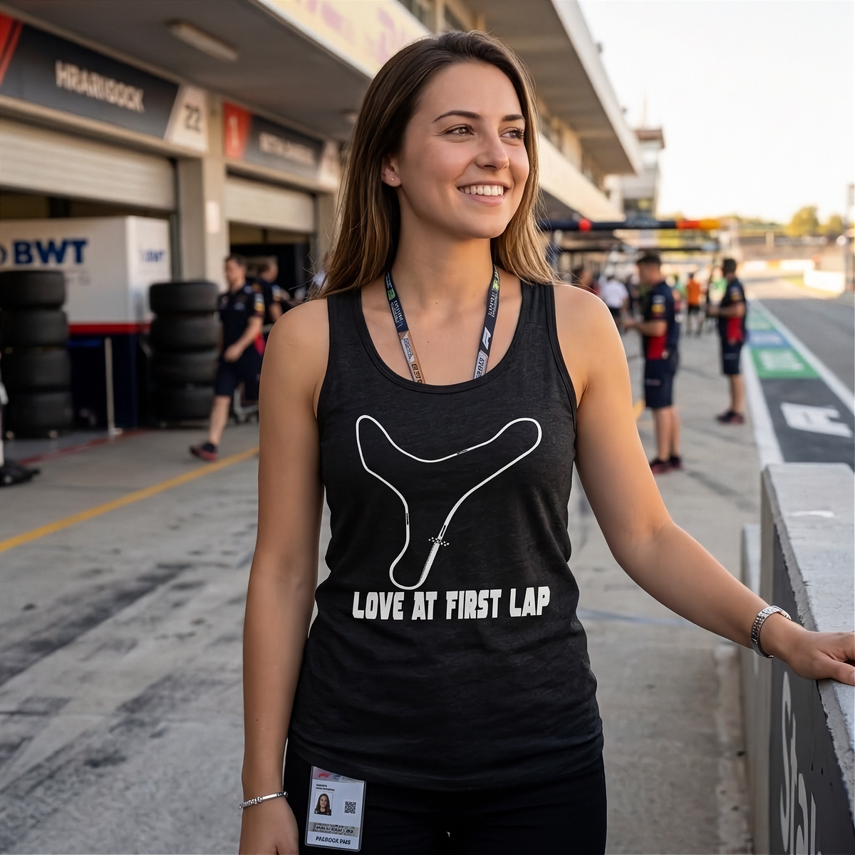 A woman standing at a racetrack pit lane wearing a black racerback tank top with a white Y-shaped track outline and the text 'LOVE AT FIRST LAP'.