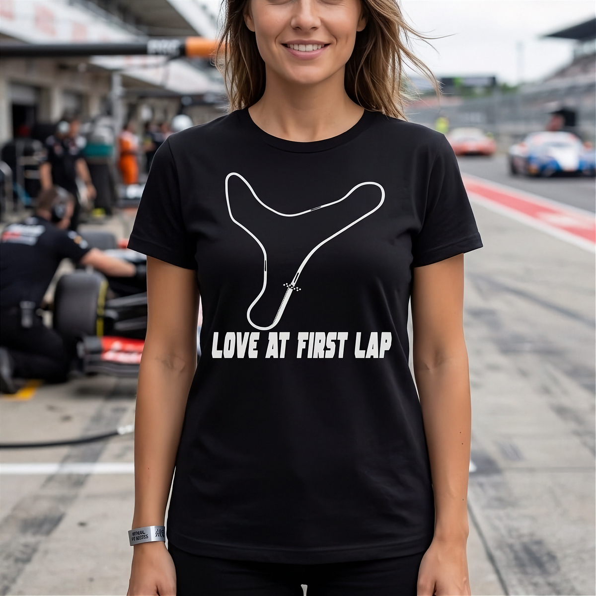 A woman wearing a black short-sleeve t-shirt with a white Y-shaped track outline and the text 'LOVE AT FIRST LAP' printed on the front, standing on a racetrack with race cars and pit crew in the background.