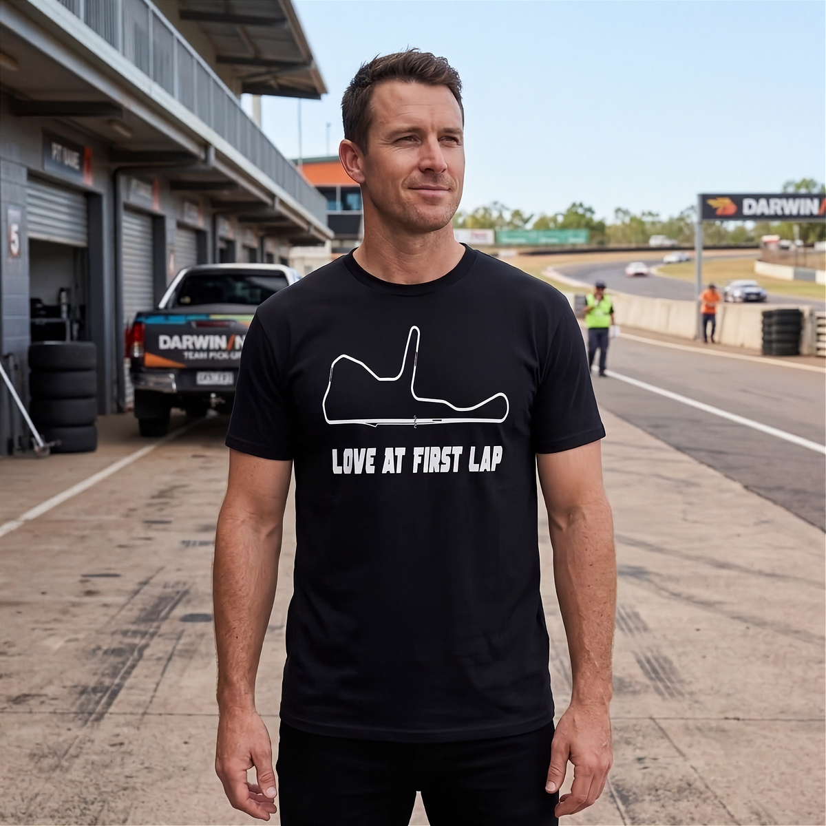 A man wearing a black crewneck t-shirt with a white racing track outline and the text 'LOVE AT FIRST LAP' stands on a racetrack paddock area. The background shows a pitlane, garage bays, and a race track under a clear sky.