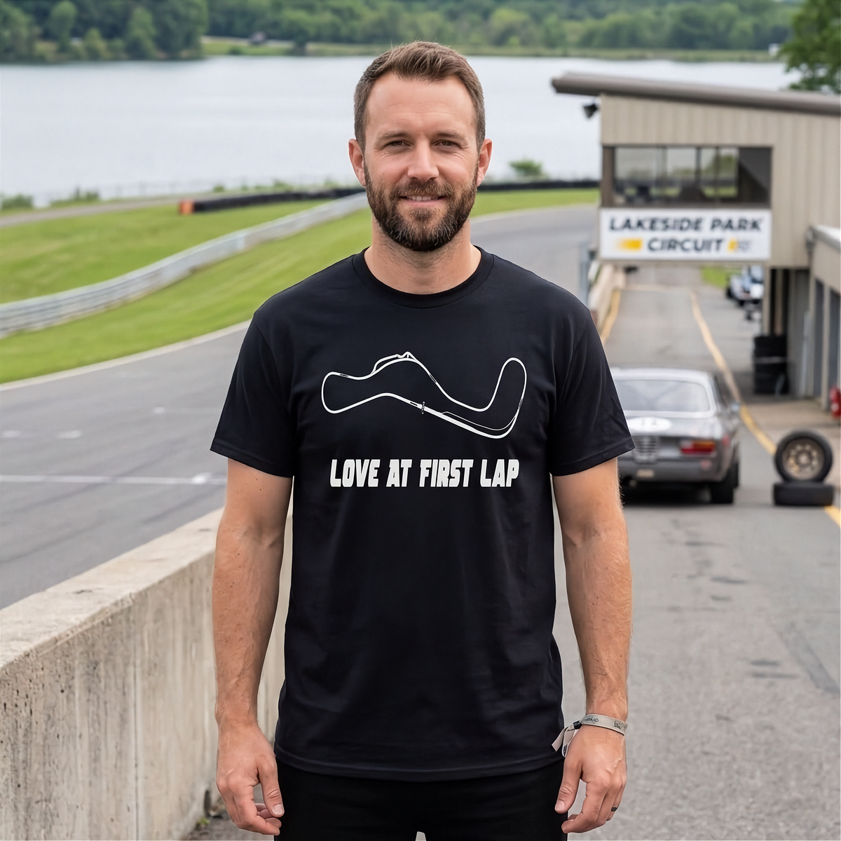 A man wearing a black t-shirt with a white graphic of a race track and the text 'LOVE AT FIRST LAP' stands on a racetrack road with a building and water in the background.