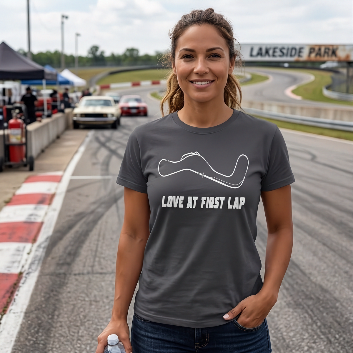 A woman stands on a racetrack wearing a dark gray t-shirt with a white racing line graphic and the text 'LOVE AT FIRST LAP'. The background shows a blurred racetrack with cars and a sign reading 'LAKESIDE PARK'.