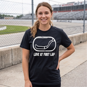 A woman smiling outdoors on a racetrack, wearing a black short-sleeve T-shirt with a white oval racing graphic and the text 'LOVE AT FIRST LAP' on the front.
