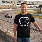 A man standing on a racetrack wearing a black t-shirt with a white track map graphic and the text 'LOVE AT FIRST LAP'. A vintage race car is visible on the track in the background under a sign that reads 'MILWAUKEE MILE SINCE 1903'.
