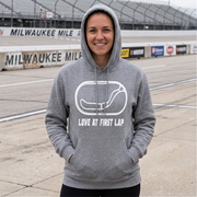 A woman wearing a heather gray pullover hoodie with a white graphic of a race car and the text 'LOVE AT FIRST LAP' stands on a racetrack with 'MILWAUKEE MILE' signage in the background.