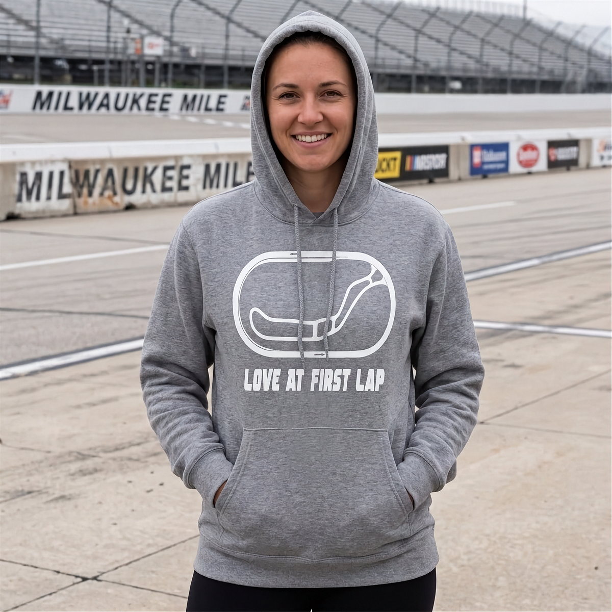 A woman wearing a heather gray pullover hoodie with a white graphic of a race car and the text 'LOVE AT FIRST LAP' stands on a racetrack with 'MILWAUKEE MILE' signage in the background.
