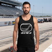 A man standing on a race track wearing a black tank top with a white graphic of a race track and the text 'LOVE MY FIRST LAP'. The background shows the racetrack, grandstands, and race cars under a clear blue sky.