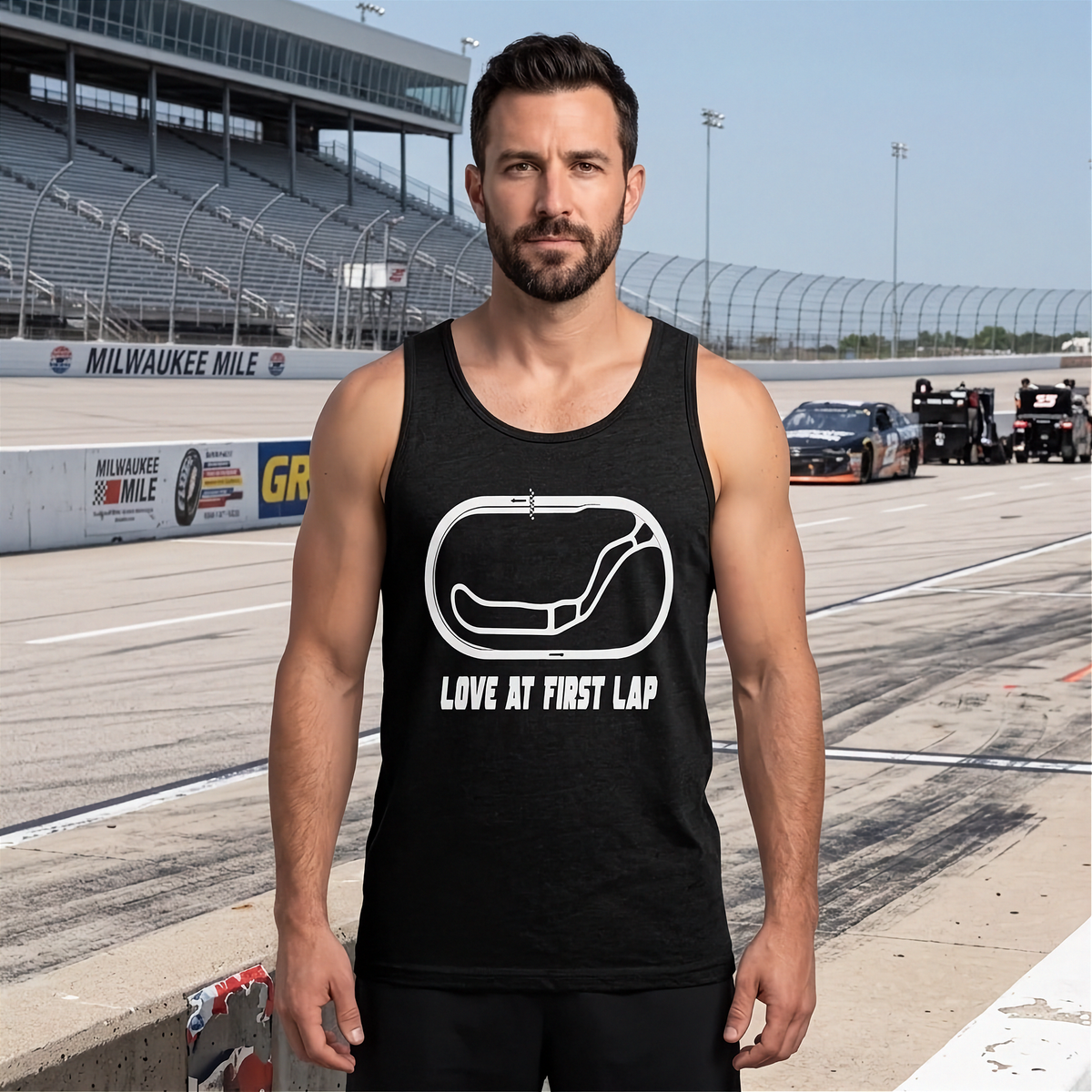 A man standing on a race track wearing a black tank top with a white graphic of a race track and the text 'LOVE MY FIRST LAP'. The background shows the racetrack, grandstands, and race cars under a clear blue sky.