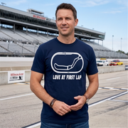 A man wearing a navy blue t-shirt with a white track graphic and the text 'LOVE AT FIRST LAP' stands on a racetrack with grandstands in the background.