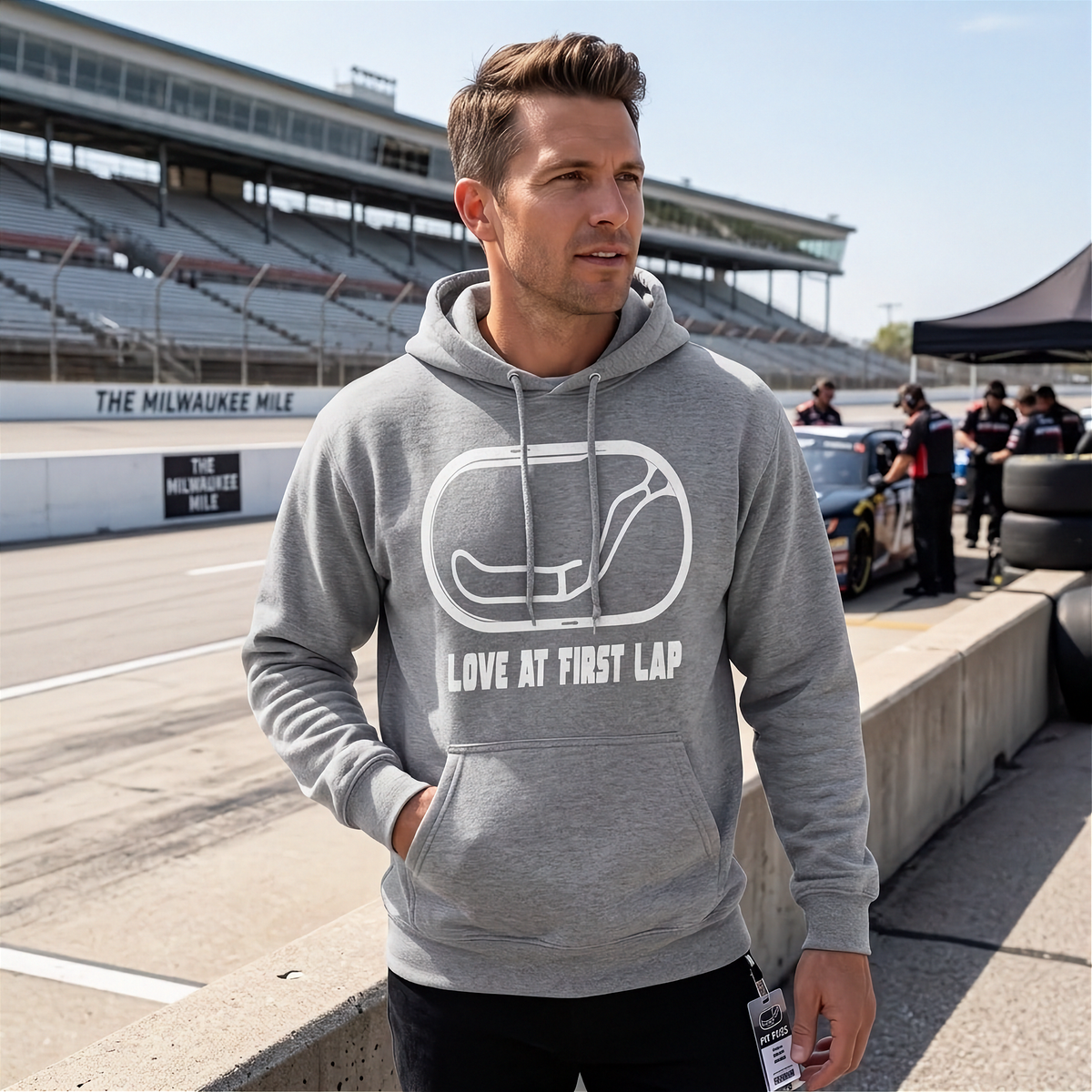 A man wearing a heather gray hoodie with a white graphic of a race track and the text 'LOVE AT FIRST LAP' stands on a racetrack. The background shows grandstands, a race car, and pit crew members.