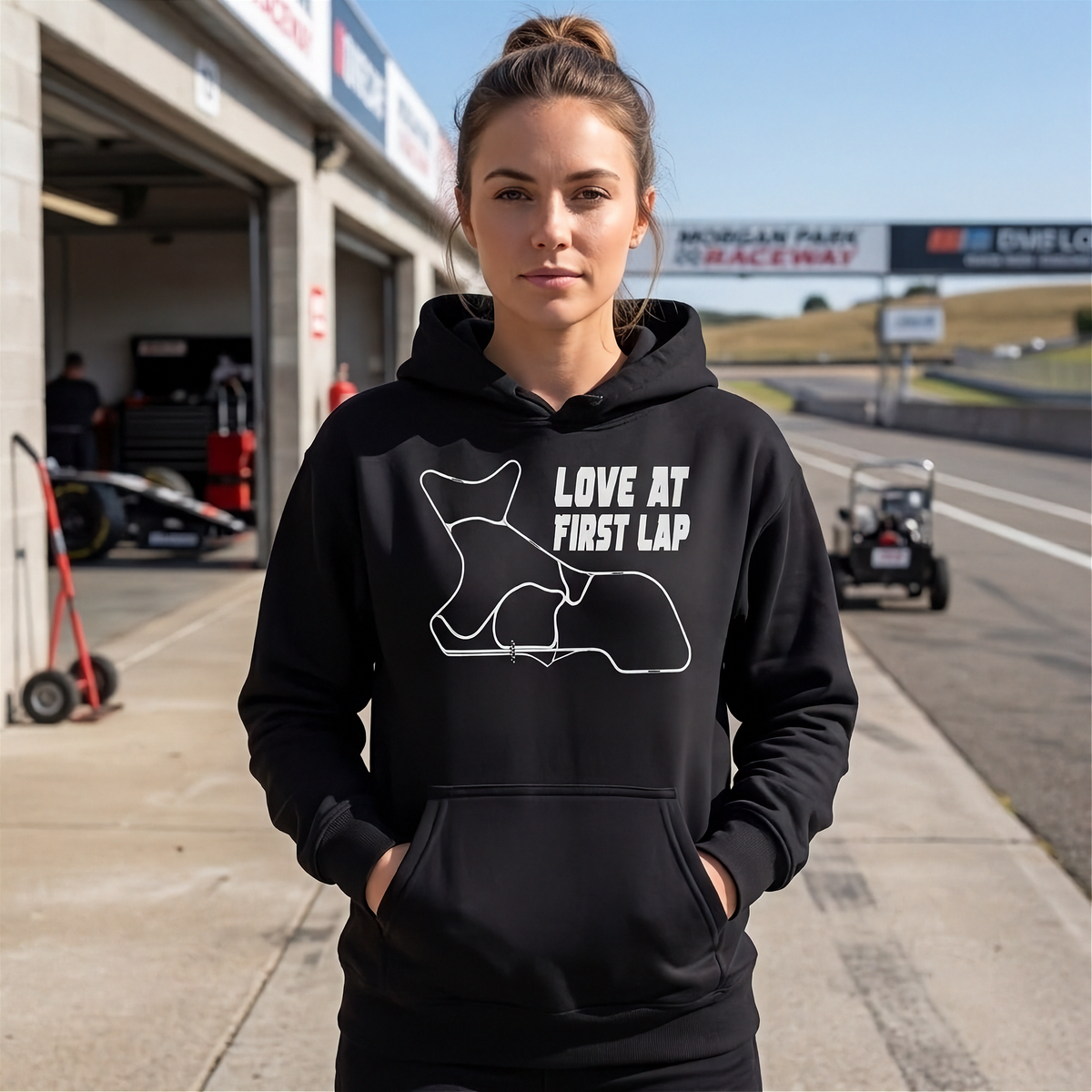 A woman stands on a racetrack wearing a black hoodie with a white outline of a race track and the text 'LOVE AT FIRST LAP'.