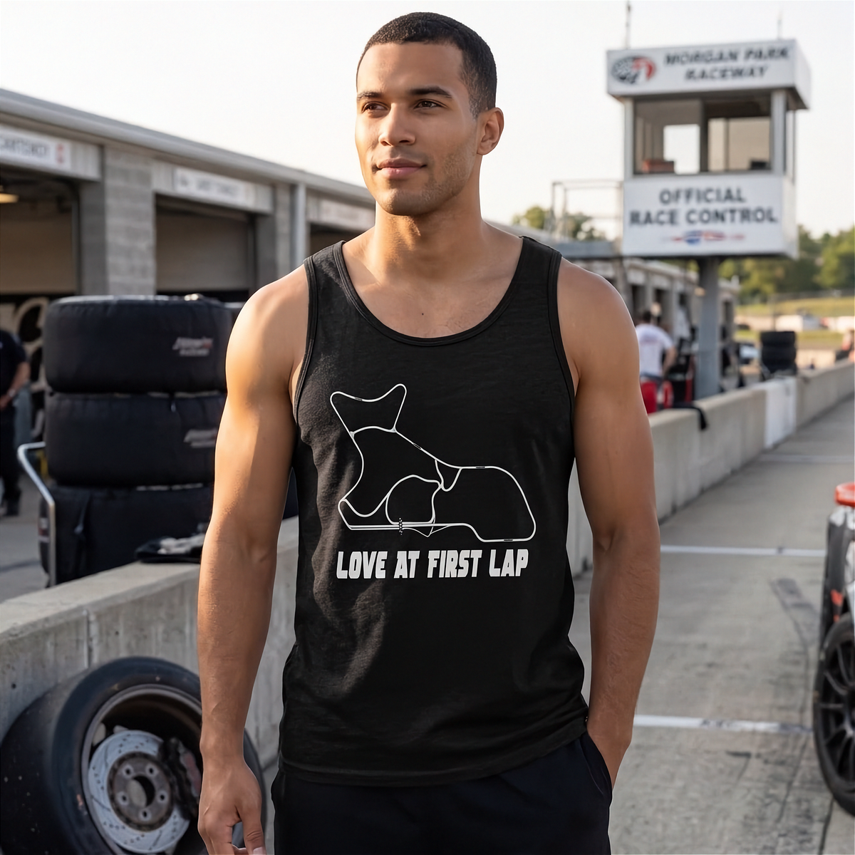 A man wearing a black tank top with a white graphic of a racing dog and the text 'LOVE AT FIRST LAP' stands at a race track pit area. The background shows race control building, tires, and a race car.
