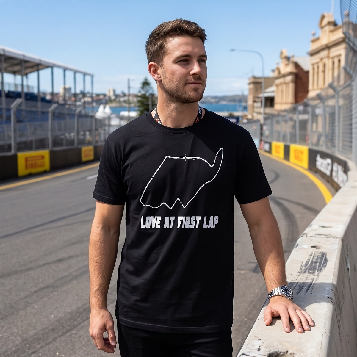 A man wearing a black t-shirt with a white track outline and the text 'LOVE AT FIRST LAP' stands on a racetrack barrier with a race course in the background.
