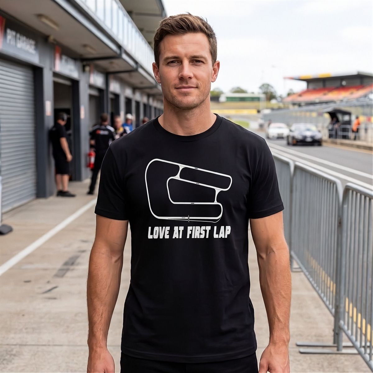 A man wearing a black t-shirt with a white racing hand graphic and the text 'LOVE AT FIRST LAP' stands on a racetrack paddock walkway.
