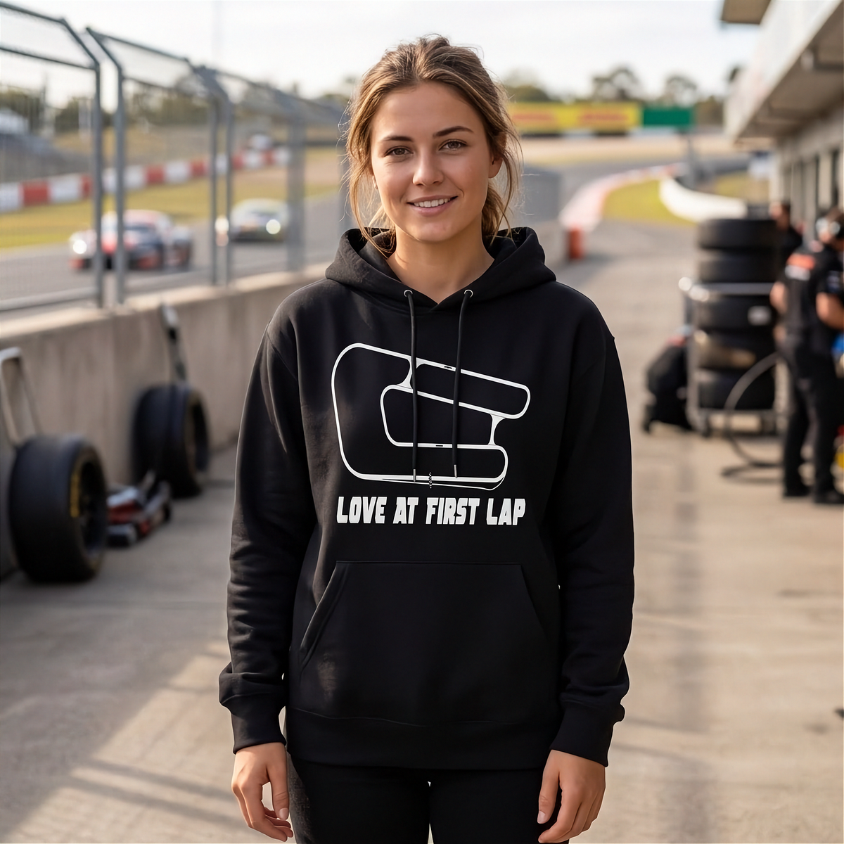 A woman stands on a racetrack wearing a black pullover hoodie with a white racing lap graphic and the text 'LOVE AT FIRST LAP' on the front.