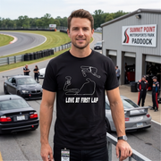 A man standing on a racetrack wearing a black t-shirt with a white line-art graphic of a race car and the text 'LOVE AT FIRST LAP'. The background shows a gray building with a sign reading 'SUMMIT POINT MOTORSPORTS PARK PADDOCK', race cars, and track staff.