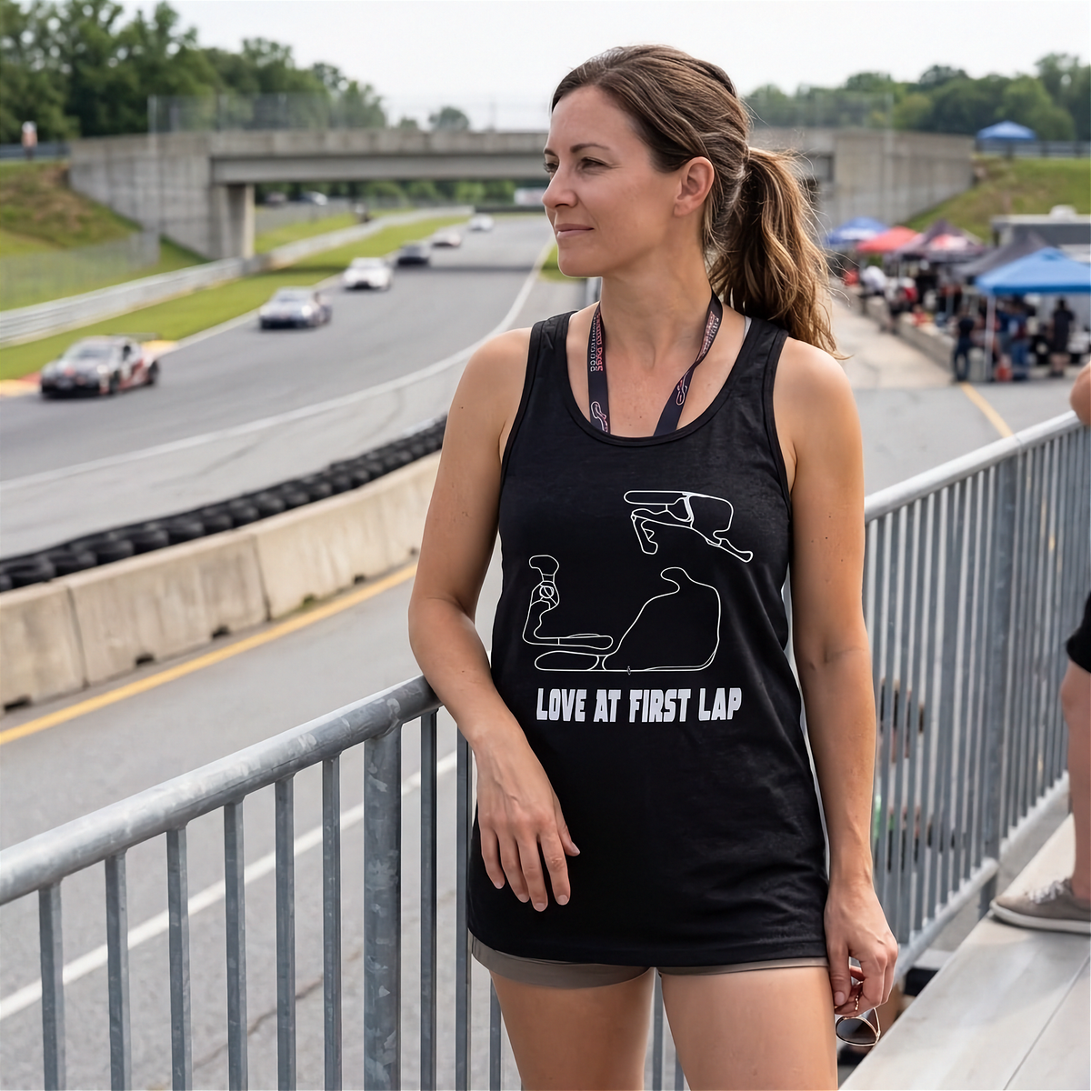 A woman stands on a bridge overlooking a racetrack, wearing a black racerback tank top with a white graphic of a race car seat and the text 'LOVE AT FIRST LAP'.