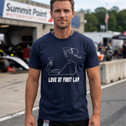 A man wearing a navy blue t-shirt with a white line art graphic of a race car and the text 'LOVE AT FIRST LAP' stands on a racetrack. The background shows a pit garage with a race car and a sign for 'Summit Point Motorsports Park'.
