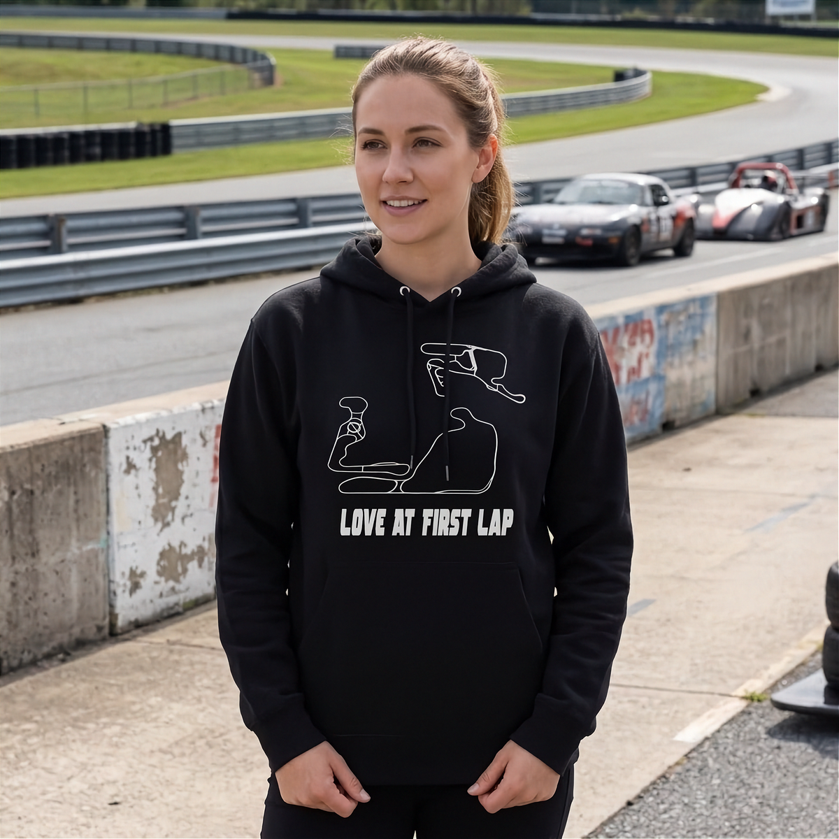 A woman wearing a black hoodie with a white graphic of a racing track and the text 'LOVE AT FIRST LAP' standing on a race track with 'SUMMIT POINT RACEWAY' in the background.