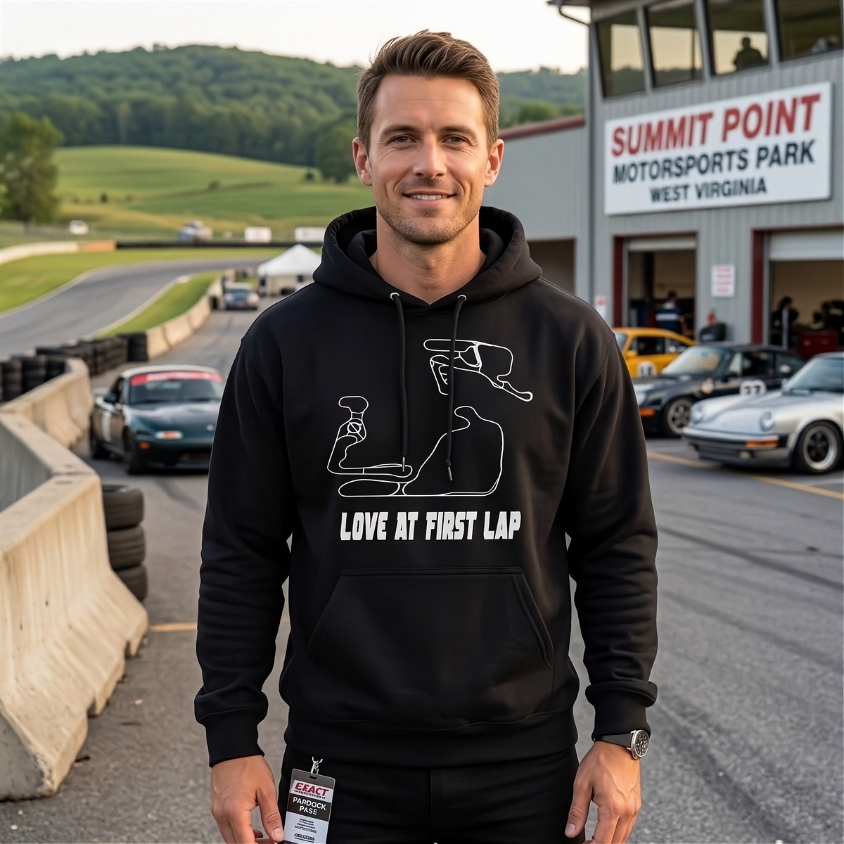 A man wearing a black pullover hoodie with a white minimalist racing line-art graphic and the text 'LOVE AT FIRST LAP' stands on a racetrack. The background shows a race track, vintage cars, and a building labeled 'SUMMIT POINT MOTORSPORTS PARK WEST VIRGINIA'.