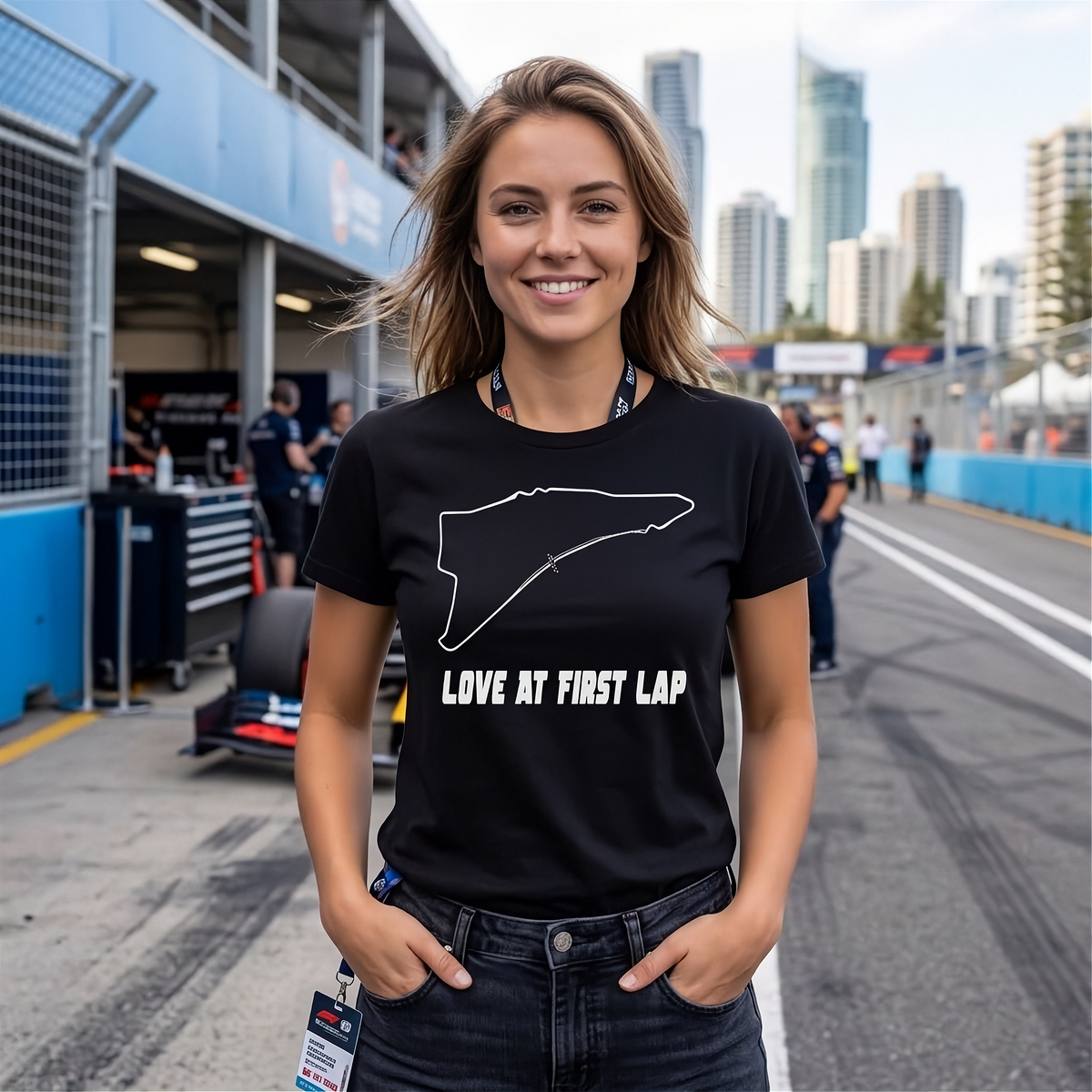 A woman stands on a race track pit area wearing a black graphic T-shirt with a white track outline and the text 'LOVE AT FIRST LAP'.
