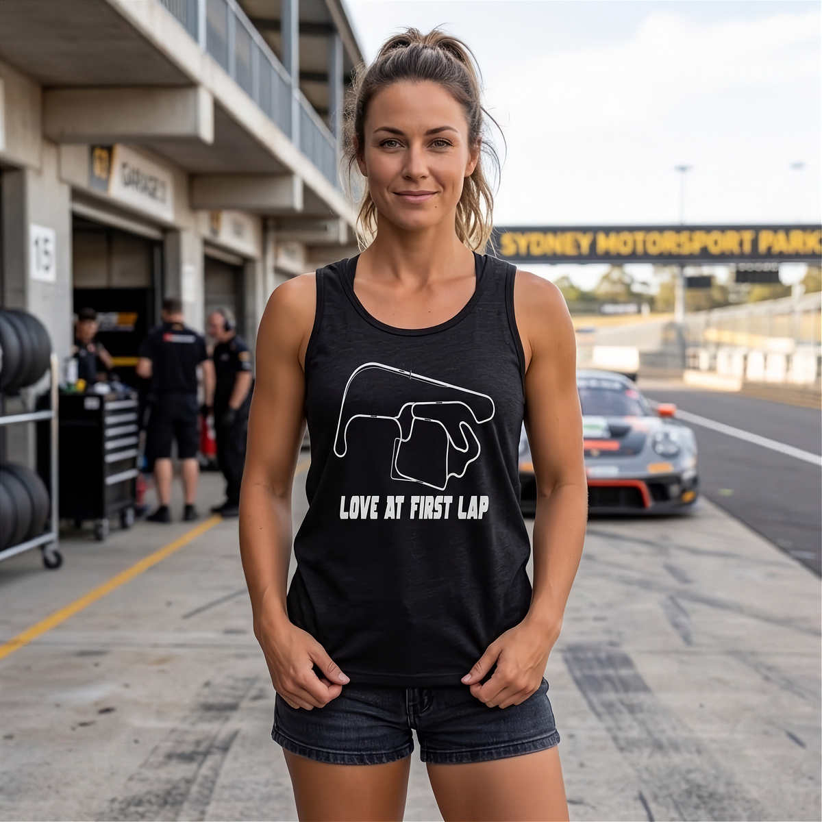 A woman stands on a racetrack pit lane wearing a black racerback tank top with a white track outline graphic and the text 'LOVE AT FIRST LAP'.