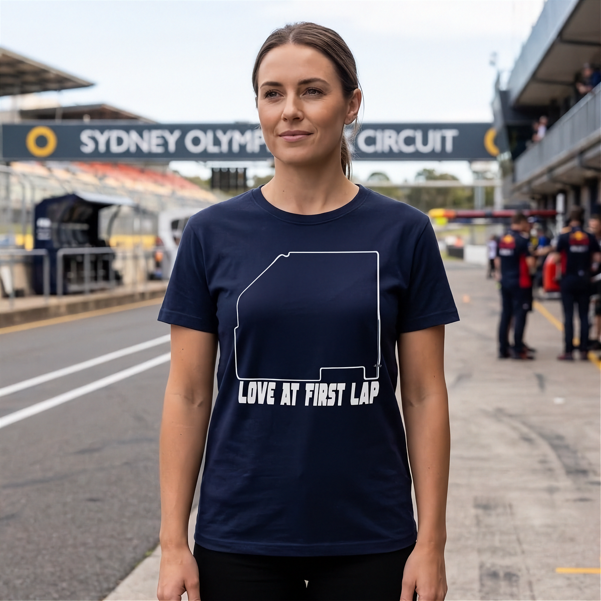 A woman stands on a racetrack wearing a navy blue t-shirt with a white outline of New South Wales and the text 'LOVE AT FIRST LAP'. The background shows the Sydney Olympic Circuit pit area.