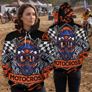 A woman modeling a black hooded sweatshirt with a large motocross helmet graphic in blue, orange, white, and black, plus checkered flag patterns and the word 'MOTOCROSS' across the front and back. The background shows an outdoor dirt track with motorcycles and tents.