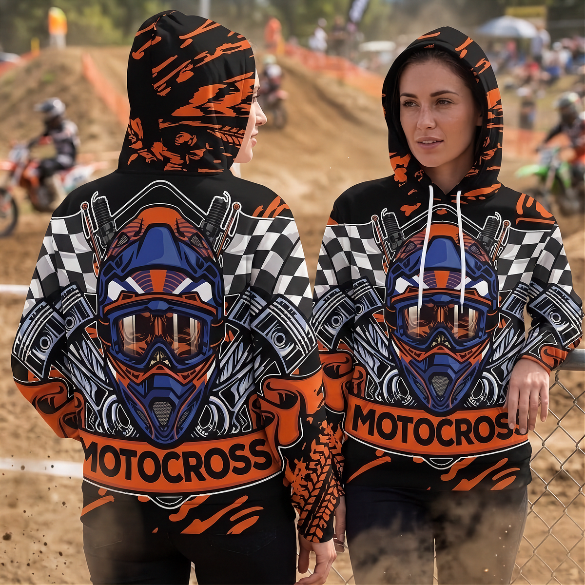 Two women modeling matching motocross hoodies at an outdoor dirt track event. The hoodies feature a large graphic of a motocross helmet and goggles on the back and front, with orange and black graphic patterns, checkered side panels, and 'MOTOCROSS' text across the waist.