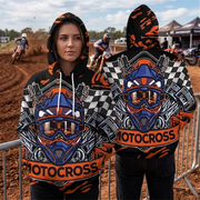A person modeling a black and orange motocross hoodie in front of a dirt track, with a second view showing the back of the same hoodie. The hoodie features a large motocross helmet graphic in blue, orange, white, and black, with checkered side panels and the word 'MOTOCROSS' printed across the front and back.