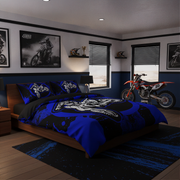 A blue and black motocross-themed bedding set with a skull graphic, styled in a modern, dark-themed bedroom featuring a dirt bike, framed wall art, and blue-and-white walls.