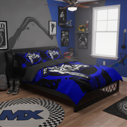 A blue, black, and white motocross-themed bedding set with matching pillowcases is displayed on a bed in a styled bedroom. The room features a gray and blue accent wall, a window, a ceiling fan, and various motocross-themed decor items including a checkered rug and framed posters.