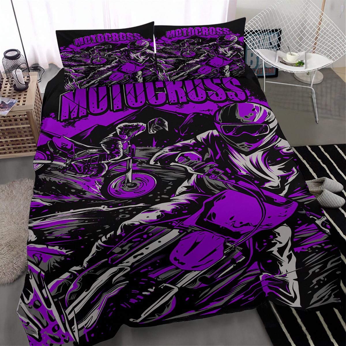 A bed dressed in a motocross-themed bedding set with a duvet cover and two pillowcases, featuring a bold graphic print in purple, black, and white of a rider on a motorcycle. The room setting includes a white wire chair, a small table with a lamp, and a striped rug.