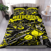 A bed dressed in a black and neon yellow motocross-themed bedding set, including a duvet cover and two pillowcases, shown in a styled bedroom with a side table, lamp, plant, and window.