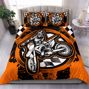 A bed dressed in a motocross-themed bedding set featuring a large graphic of a rider on a dirt bike over a black-and-orange splatter design with checkered accents. Two matching pillowcases are placed behind the duvet.