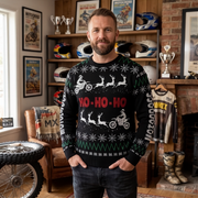 A man wearing a black Christmas sweater with a red and white motocross theme stands in a rustic room with motorcycle helmets, framed posters, and trophies in the background.