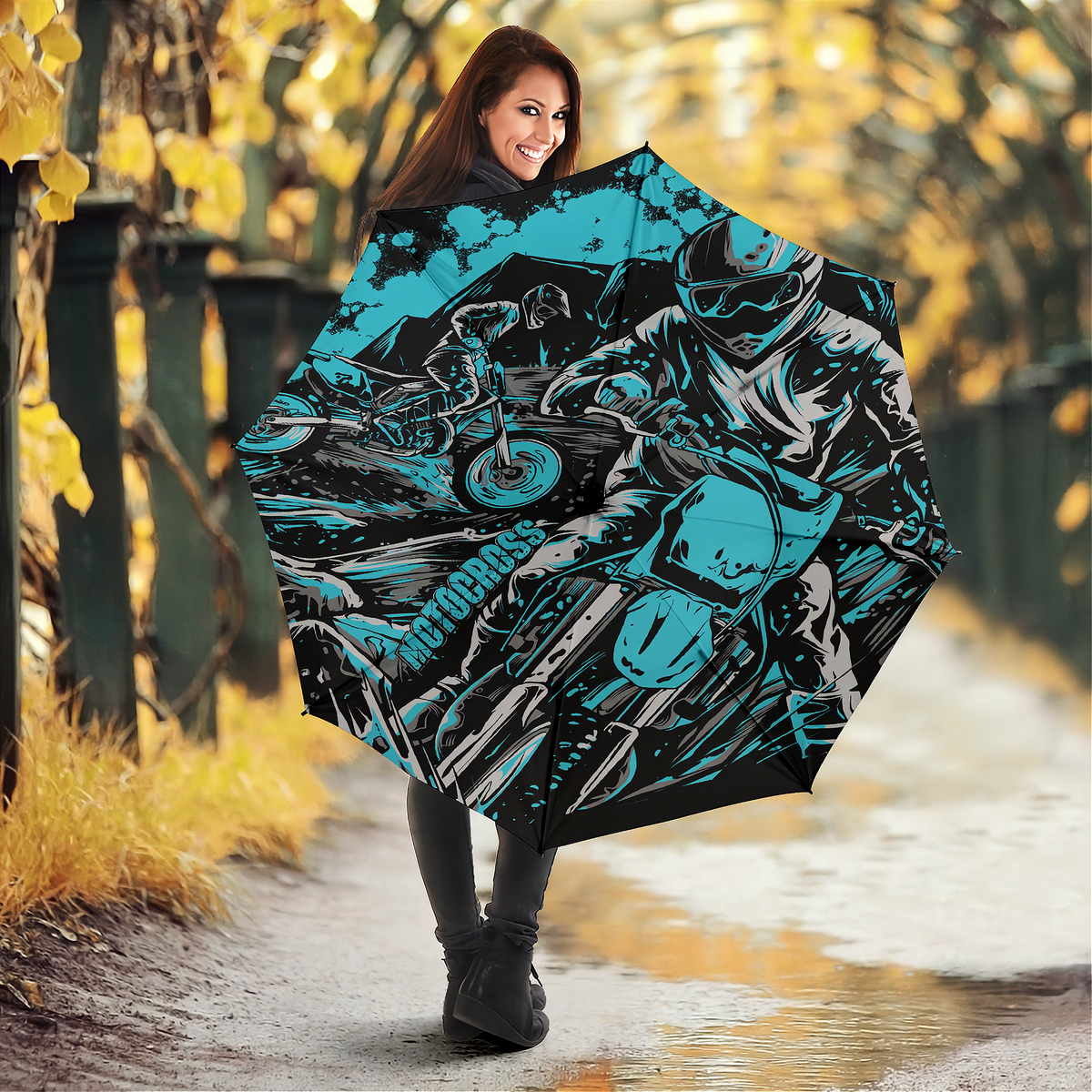 A woman holding an open umbrella in an outdoor autumn setting. The umbrella features a bold graphic print of a motorcyclist in Carolina Blue, black, and white, with the word 'Motocross' printed vertically on the side.