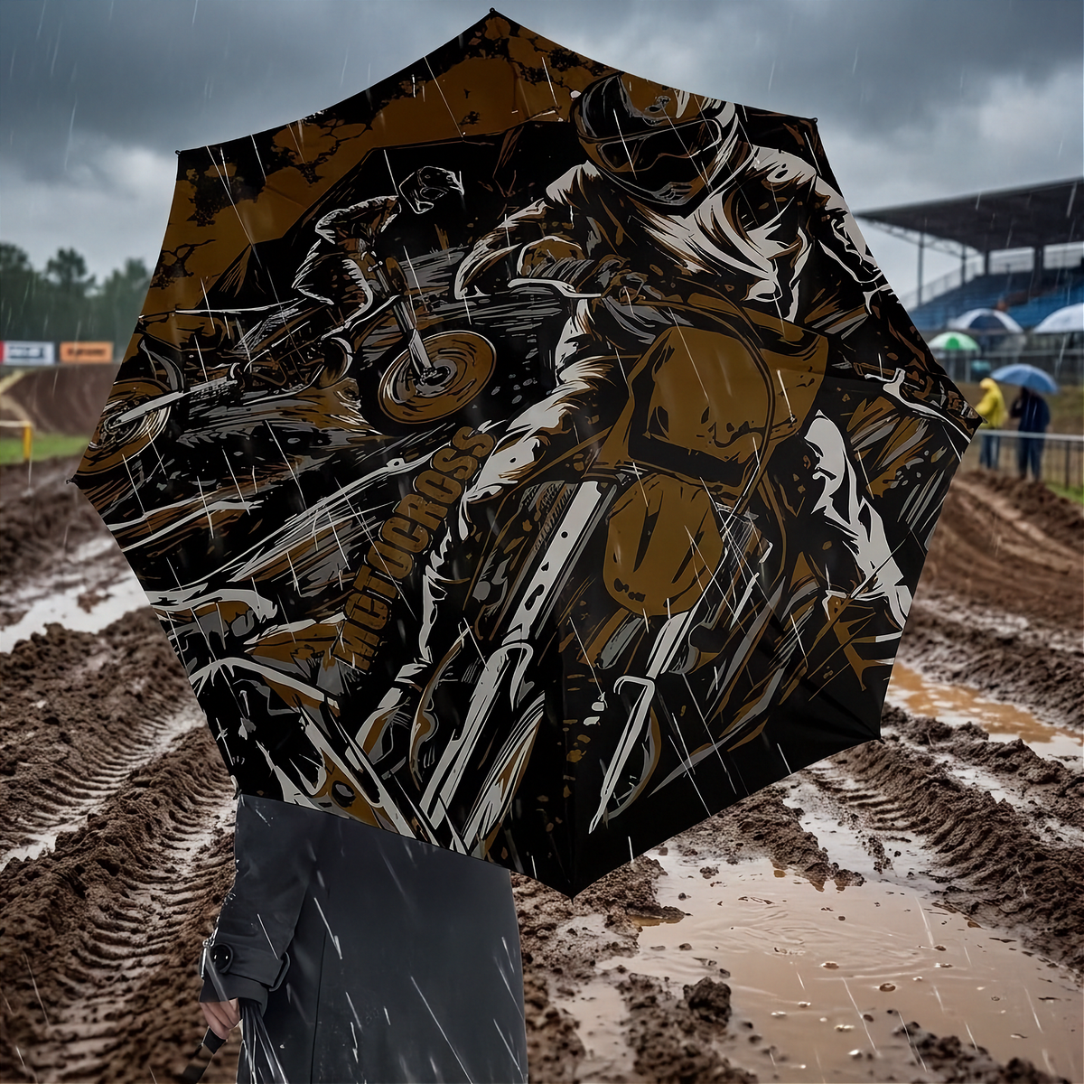 An open octagonal umbrella with a bold brown and black motocross graphic print, held up in the rain over a muddy outdoor track.