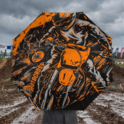 An open octagonal umbrella with a bold orange and black motocross graphic print, held up in the rain over a muddy outdoor track.