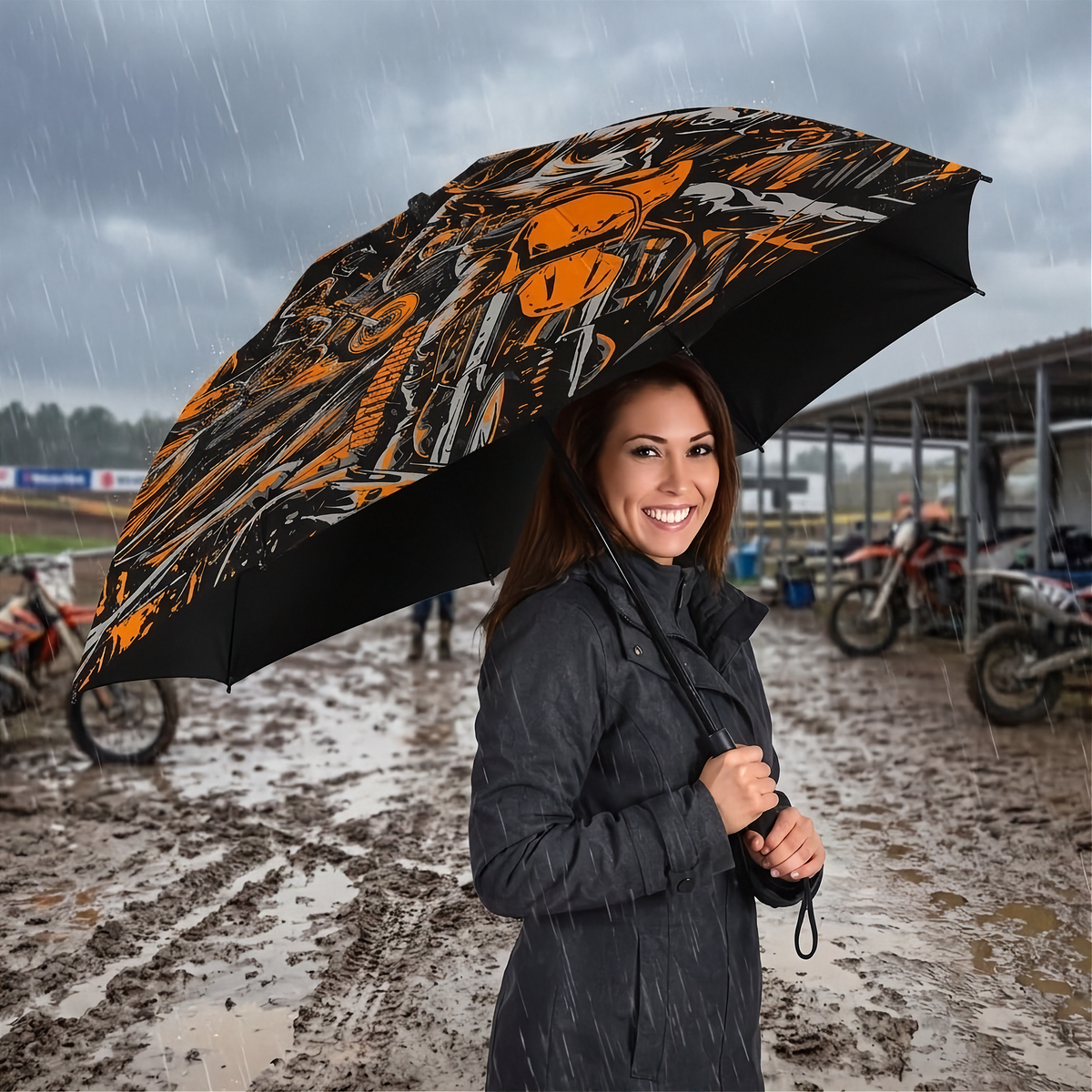 A woman in a dark coat stands in the rain holding a large open umbrella with a bold orange, black, and white graphic pattern. The background shows a muddy outdoor dirt bike racing.