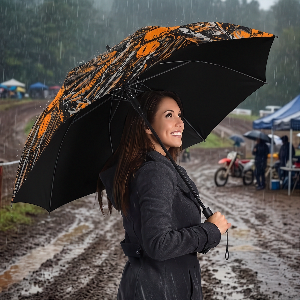 A woman in a dark gray raincoat stands in the rain holding a large open umbrella with a bold orange, black, and white graphic pattern. The background shows a muddy outdoor path and a cloudy sky.