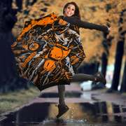 A woman jumps in a puddle outdoors while holding open a large umbrella printed with a bold orange, black, and gray motorcycle graphic. Trees with yellow autumn foliage are blurred in the background.