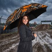 A woman in a dark raincoat stands in the rain at a muddy motocross track holding an open umbrella with a black background and a orange, black, and white graphic pattern.