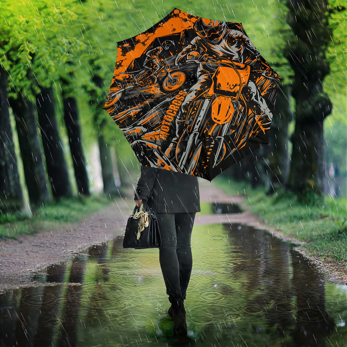 A person walking in the rain on a tree-lined path holding a large open umbrella with a orange, black, and white motorcycle graphic print.