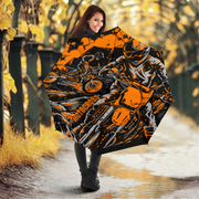 A woman holding an open umbrella in an outdoor autumn setting. The umbrella features a bold graphic print of a motorcyclist in orange, black, and white, with the word 'Motocross' printed vertically on the side.