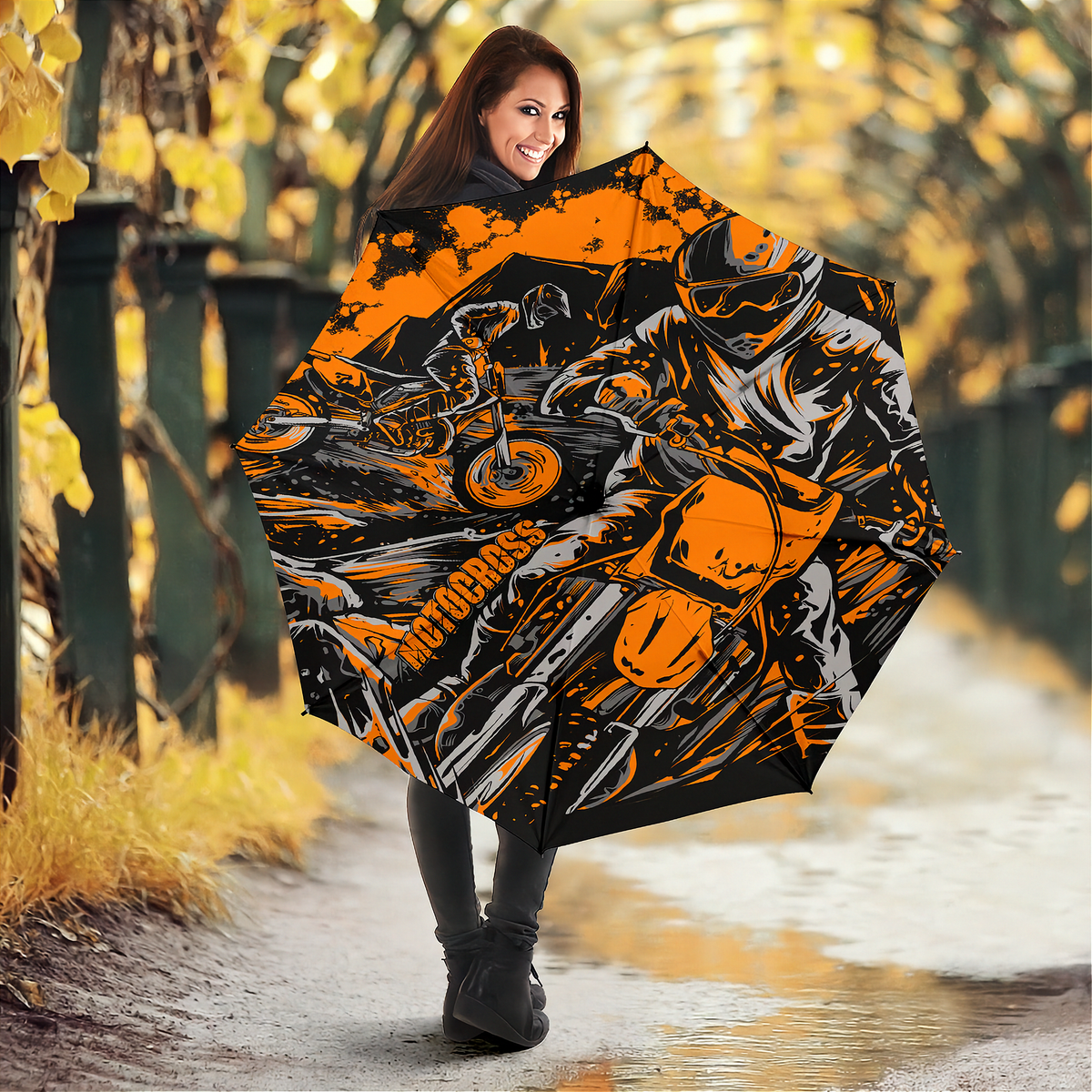 A woman holding an open umbrella in an outdoor autumn setting. The umbrella features a bold graphic print of a motorcyclist in orange, black, and white, with the word 'Motocross' printed vertically on the side.