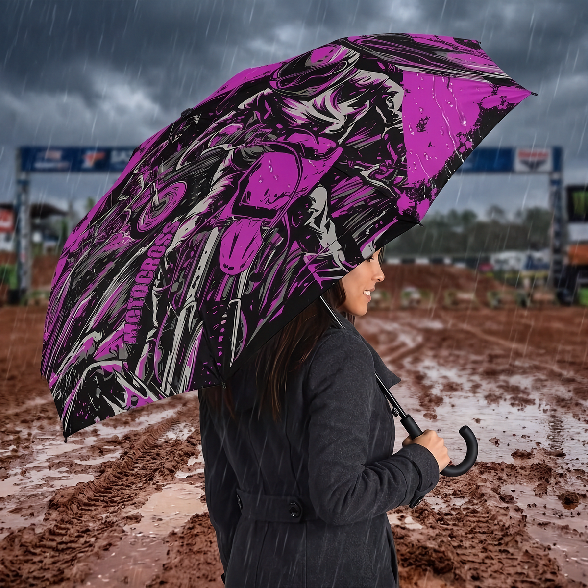 A woman in a dark coat stands in the rain holding a large open umbrella with a bold pink, black, and white graphic pattern. The background shows a muddy outdoor event or track with banners and blurred figures.