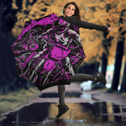 A woman jumps in a puddle outdoors while holding open a large umbrella printed with a bold pink, black, and gray motorcycle graphic. Trees with yellow autumn foliage are blurred in the background.