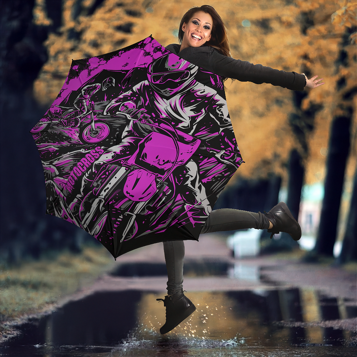 A woman jumps in a puddle outdoors while holding open a large umbrella printed with a bold pink, black, and gray motorcycle graphic. Trees with yellow autumn foliage are blurred in the background.